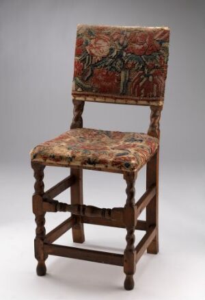  Antique wooden chair with turned spindle legs and horizontal stretchers, and richly patterned upholstery in red, green, and gold tones on both the seat and backrest, set against a neutral gray background.