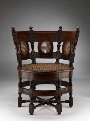  An antique dark brown wooden chair with a caned seat and two caned circular panels on the backrest, decorated with turned wood details, spherical finials, and intricate carvings, against a grey backdrop.