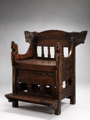  Traditional wooden chair, known as Gårastolen, made of pine and birch with a rich brown color. It features a sturdy post construction with a high back decorated with carved stylized animal head or scroll designs at the corners, vertical slats, and a lower footrest platform, manifesting Scandinavian or Norwegian folk furniture craftsmanship. Artist unknown.