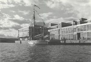  Black and white photograph of a modern, geometric multi-level building next to a calm body of water with a traditional ship docked alongside, under a partly cloudy sky.