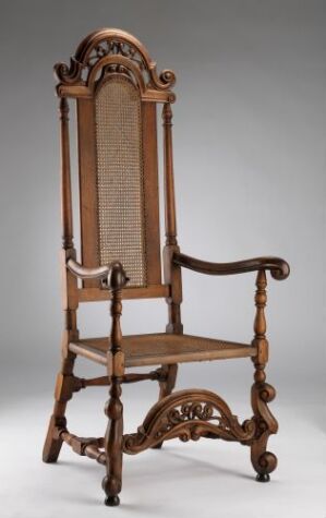  An antique wooden chair with a high backrest featuring woven cane, intricate carvings and turned spindles, all in a rich warm brown hue against a subdued gray background.