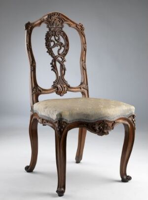  An ornate vintage wooden chair with rich brown glossy finish and creamy upholstered seat, featuring intricate carvings of organic motifs on the backrest and legs, set against a neutral grey background.