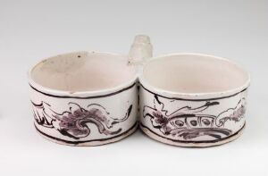  A Herrebøe Fajansefabrikk double container made of white-glazed faience with hand-painted manganese red decorations, featuring two joined circular sections without lids and a connecting handle, displaying intricate patterns.