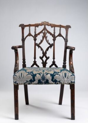  An antique wooden armchair with detailed gothic-style lattice backrest and an upholstered seat with a complex blue, gray, and off-white pastoral scene, set against a light gray background.