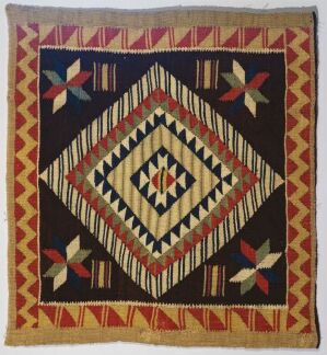  A square quilt with a black background and a symmetrical pattern featuring a series of concentric diamonds in white, yellow, red, green, and blue colors, with corner flower-like motifs and a red and yellow chevron border.