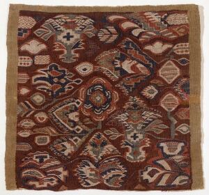  Square woven textile with a symmetrical pattern in earth tones, featuring a central octagonal medallion surrounded by geometric and floral designs, enclosed by a patterned border.