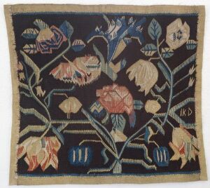  A textile artwork with a dark background featuring stylized plants and flowers in muted colors including pale yellow, beige, ivory, pink, coral, green, blue, and touches of red. The image shows the tapestry with slightly frayed edges and the initials "K.D." in the lower right corner.