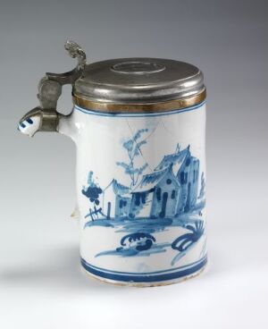  A blue and white ceramic tankard with a hinged metal lid, depicting a rustic European scene with a building and vegetation.