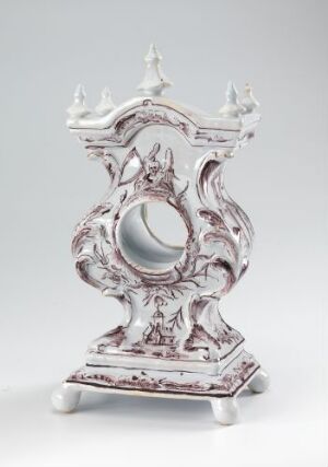  A ceramic piece titled "Uhrværk-Casse" by Herrebøe Fajansefabrikk, showcasing a white-glazed earthenware with hand-painted manganese red decor. The design is ornate with baroque influences, featuring a central circular opening, possibly for a clock face, with curvaceous edges and a sturdy rectangular base. The intricate floral motifs and swirling patterns convey an antique charm.