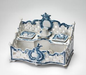  A rectangular white ceramic inkstand with scalloped edges and ornate blue floral patterns. It features two square containers with lids and a central round holder, all adorned in matching blue and white design, and an ornamental blue piece resembling a crest at the center back of the stand.