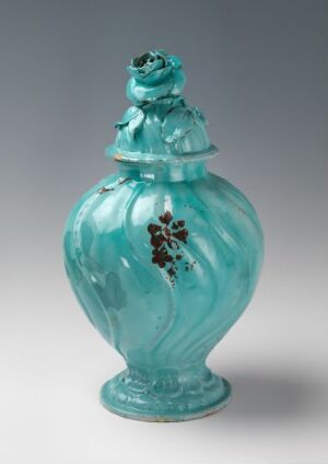  A bulbous turquoise-green faience vase with a rounded body and a decorative lid featuring remnants of handpainted gold floral motifs. The vase has a glossy finish and sits against a neutral background. Artist name and title are unknown.