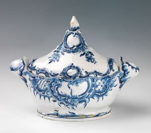  A white glazed faience tableware piece with intricate hand-painted cobalt blue designs, featuring floral patterns and scrolled embellishments, suggestive of baroque or rococo styles. The item has a rounded base, a curvaceous body, and a domed lid with a decorative finial. Artist name unknown.