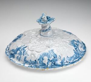  A white ceramic lid with blue floral and leaf patterns and an ornate, wavy edge, topped with a small blue-and-white handle in the center, displayed on a grey background.