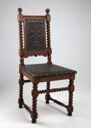  An antique dark brown wooden chair with intricate carvings on the backrest and ornamental turned spindles connecting the legs, set against a light grey background.