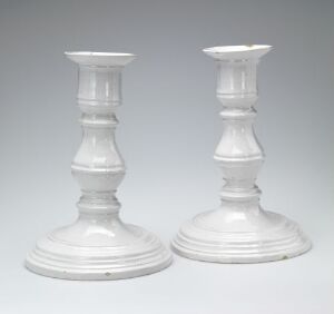  A pair of symmetrical, clear glass or crystal candlesticks with a classic design, consisting of a circular base and a decorative column, set against a light grey gradient background. Artistname and title are unknown.