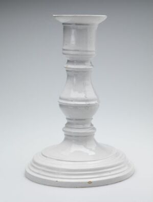  A traditional white ceramic candlestick holder with a tiered base and top set against a light grey background. The holder features a bulbous midsection and an overall elegant, timeless design.