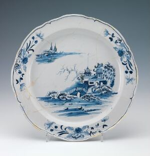  A white-glazed fajanse plate with hand-painted cobalt blue decorations, featuring a central landscape scene with a building and a sailboat, surrounded by a decorative border with floral and foliage motifs.