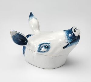  A white glazed faience tableware piece with a hand-painted cobalt blue decoration, modeled in the shape of a cow or bull's head, indicative of a whimsical serving dish or container. The piece exudes a playfulness through its expressive blue eyes and detailed features, set against a pristine white background. Artist name and title are unknown.