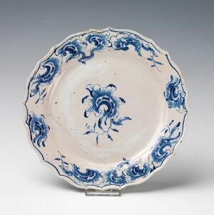  An antique, handcrafted fajanse plate with scalloped edges, featuring hand-painted cobalt blue floral designs on a white glossy background, created by an unknown artist.