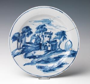  A round ceramic plate painted with a blue and white pastoral scene featuring a central house, surrounded by stylized trees and set against a hilly backdrop.