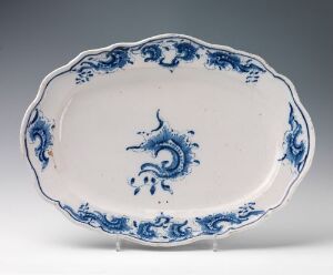  An oval-shaped, white glazed stoneware platter with scalloped edges, hand-painted with cobalt blue decorative motifs, designed by Herrebøe Fajansefabrikk. The platter features a central swirling design flanked by intricate scrollwork and floral patterns around the rim.