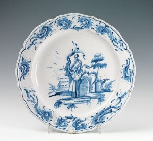  A glazed faience plate with scalloped edges, featuring handpainted cobalt blue pastoral designs with a central figure surrounded by trees, indicating traditional craftsmanship. Artist unknown.