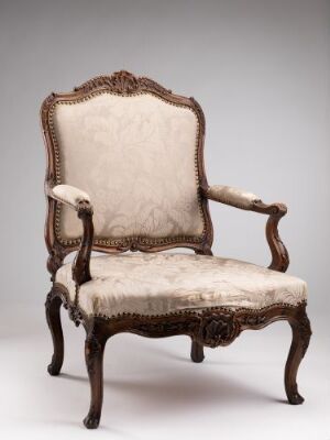  An antique, richly carved wood armchair with pale beige upholstery and brass tacks, set against a light grey background.