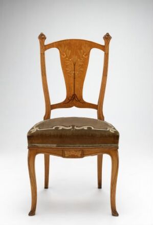  An antique wooden chair with a polished honey-brown frame and ornate carvings on the tall backrest, featuring a faded cushioned seat with a scalloped edge trim, set against a light gray background.