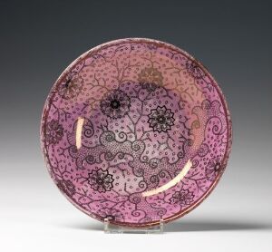  An intricately designed circular ceramic plate with a complex pink and dark ink floral pattern, featuring visible repairs in contrasting color, displayed against a plain gray background.