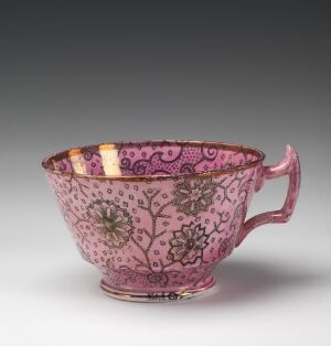  An ornate deep pink porcelain teacup with a floral and paisley pattern in darker shades, featuring a band of intricate design with touches of yellow and blue inside the rim, and a delicate gold edge at the top.