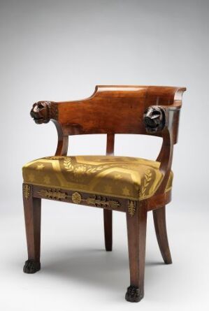  An antique chair with a curving mahogany back and arms ending in carved lion's heads, with a plush golden-yellow seat cushion and decorative gold trim just below the seat.