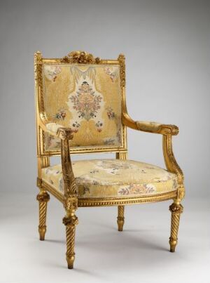  An antique gilded armchair with ornate carvings and cream upholstery featuring a symmetrical floral pattern on the backrest and seat. The chair has elegantly curved armrests and fluted legs with pointed feet, exuding an air of historical luxury.