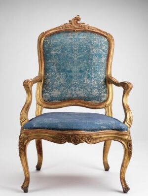  An ornate, classic chair with golden yellow-brown wooden frame, detailed with acanthus leaf carvings, and upholstered with deep blue velvet featuring subtle darker blue floral patterns, set against a white background.