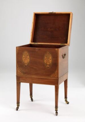  An antique wooden cabinet on tapered legs with metallic caps and casters, featuring an open top lid and two decorative inlay circles on the front, set against a neutral backdrop.