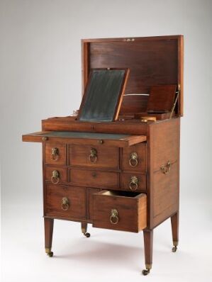  A mahogany "Toalettmøbel med skriveplate for reise" by an unidentified producer, featuring a writing surface and multiple drawers with brass fittings, set against a simple, light-colored background.