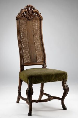  An antique "Stol" chair made from walnut (nøttetre), beech, and oak, with intricate wood carvings and a tightly woven rattan back, featuring a padded seat covered in moss green plush fabric. Artist unknown.