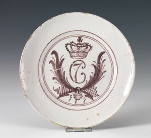  Antique cream-colored ceramic plate with a burgundy monogram, flanked by laurel branches, and topped with a crown. The plate shows signs of wear with minor chips on the rim.