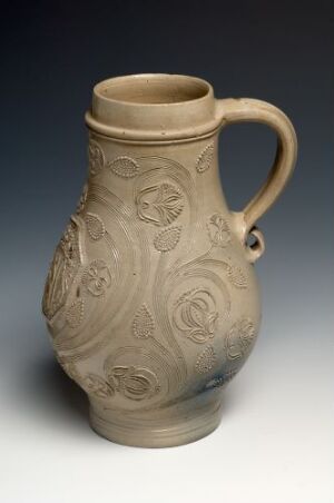  An unglazed beige ceramic pitcher with embossed floral designs and an elegantly curved handle, set against a graduated blue-gray background.