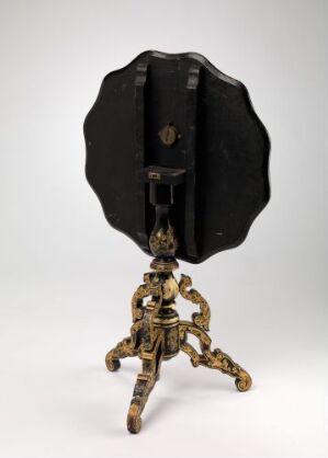  An antique bellows camera mounted on an ornate brass tripod stand against a light gray background. The camera is black with a circular front lens board and the three-legged stand features intricate scroll designs.