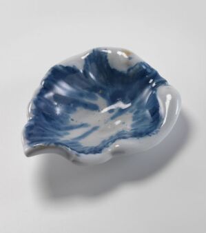  A white-glazed faience dish with hand-painted decorations in cobalt blue, titled "Complet Plat des Menager med Behør" by Herrebøe Fajansefabrikk, featuring an organic, leaf-like shape with wavy edges and a marbled blue pattern that mimics the natural flow of water.