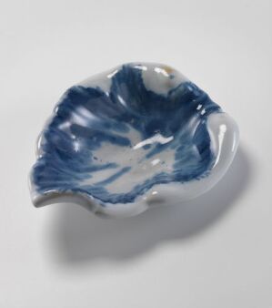  A white-glazed fajanse dish with hand-painted cobalt blue decoration in a fluid, marbled pattern, resembling a sea shell or flower petal, made by Herrebøe Fajansefabrikk. The dish has an organic, wavy edge and a richly painted interior that contrasts with the mostly white exterior.