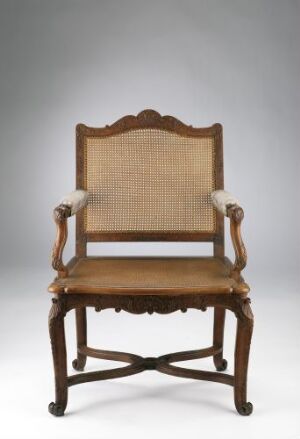  An antique wooden armchair with scrolled armrests and a tan upholstered backrest and seat, set against a plain gray background.