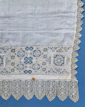  A segment of a white or off-white delicate textile with intricate lace detailing along the border, displayed over a deep sky blue background. The lacework features geometric and floral patterns with a scalloped fringe edge, and there is a small light brown stain on the fabric.