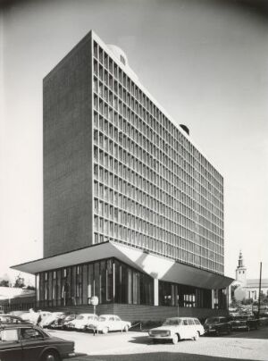 
 A black and white architectural photograph of a mid-20th century modernist building with a tall rectangular structure and a smaller base with a sloped roof, surrounded by vintage cars, under a clear sky.