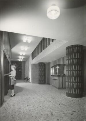  A black and white architectural photograph displaying a modern interior with a patterned carpet, a woman standing to the left reaching out towards the wall, a cylindrical bookshelf on the right, and a wide, illuminated staircase ascending diagonally to the upper level. The atmosphere conveys a sense of contemporary design and serenity.