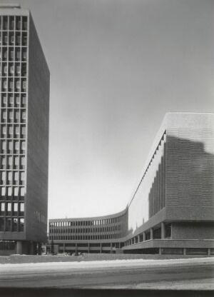  Black and white photograph of a modernist architectural complex with a tall, slender high-rise building to the left and a curved, horizontal structure to the right, under a clear sky.