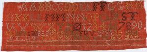  A photograph of an ancient-looking, narrow red textile band with rows of intricate white or light-colored symbolic characters or alphabetical motifs repeated along its length, showing slight fraying at the edges.