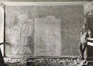 "Black and white photograph of a person standing to the right, observing two large etchings or drawings on a wall, reflecting the historical significance and artistic details within an architectural context on photographic paper."