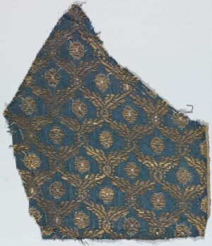  Fragment of an aged textile with an intricate golden-yellow floral pattern on a dark blue background, showing signs of wear and an irregular, almost triangular shape.