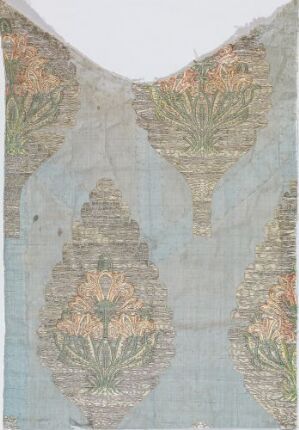  A faded blue-grey textile fragment with intricate, symmetrical embroidery in muted earthy tones, featuring a central column-like pattern flanked by similar motifs, and visibly aged with frayed edges and a V-shaped neckline cutout.